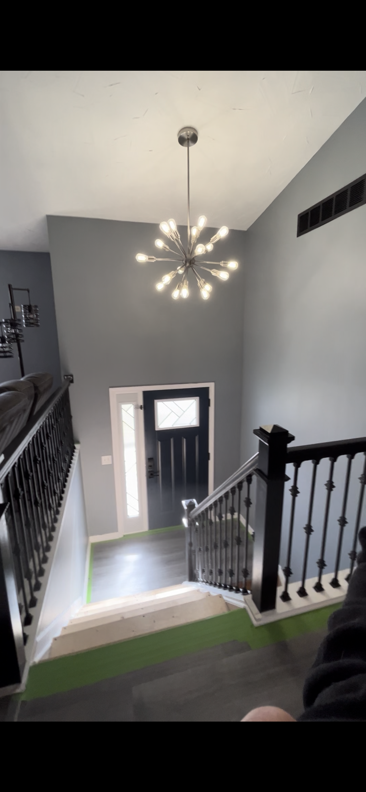 Interior view of a modern home entryway showing a staircase with black spindles and new flooring, a black front door with windows, gray walls, and a contemporary chandelier hanging from the ceiling.