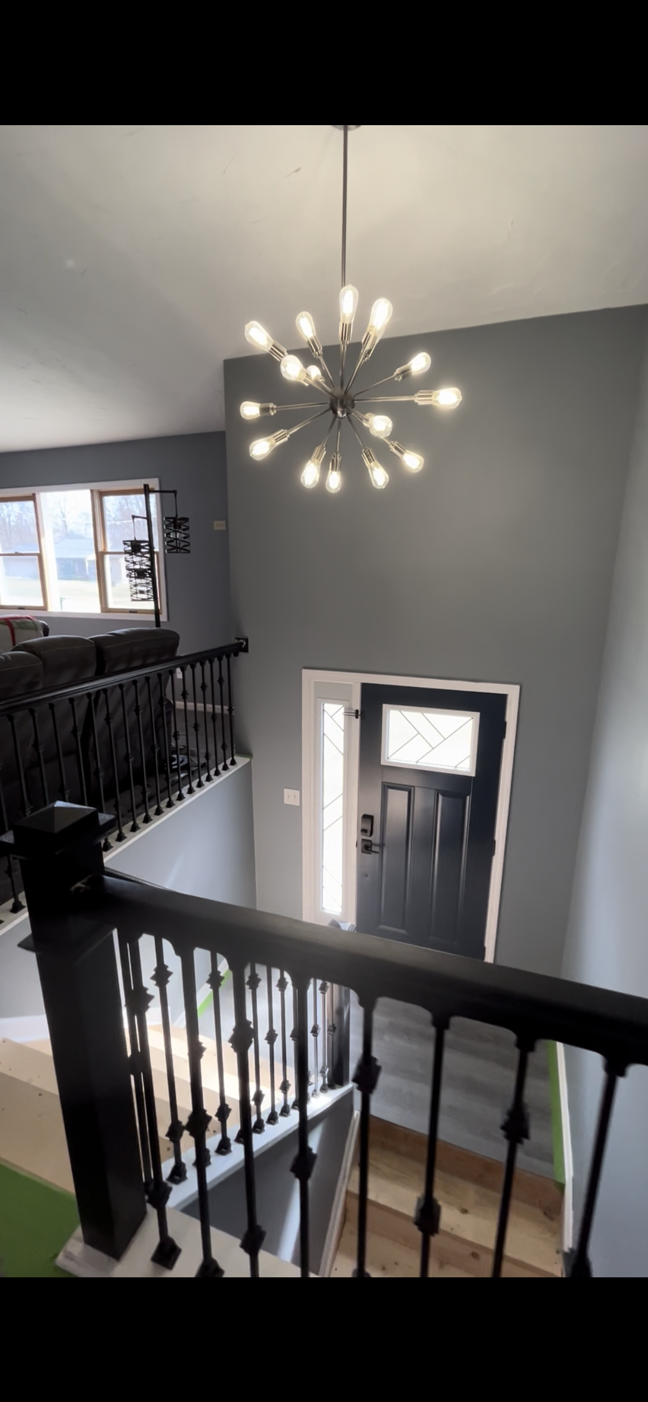 Interior view of a modern home entrance with black front door, large window, staircase with black iron railing, and a contemporary chandelier with multiple exposed bulbs hanging from the ceiling.