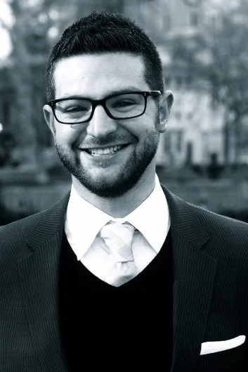 Young man in a suit, tie, and glasses smiling outdoors in black and white.