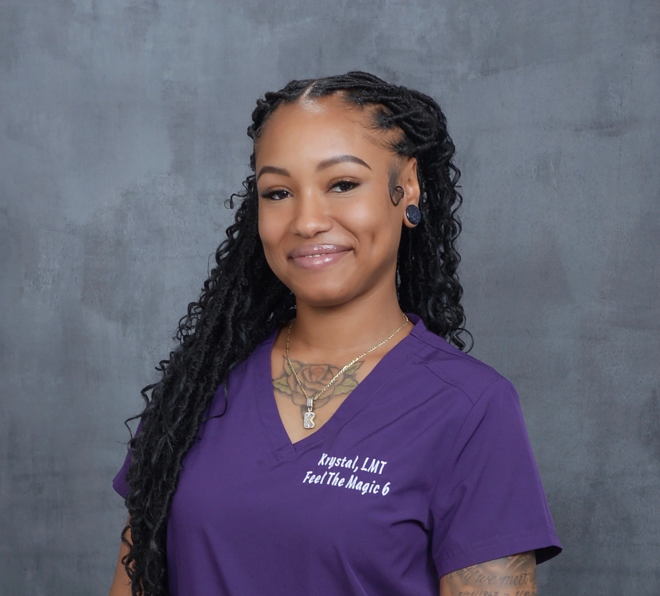 A woman with dark, curly hair styled in twists, wearing purple scrubs with name embroidery, jewelry including necklaces and earrings, standing against a gray background, smiling softly.