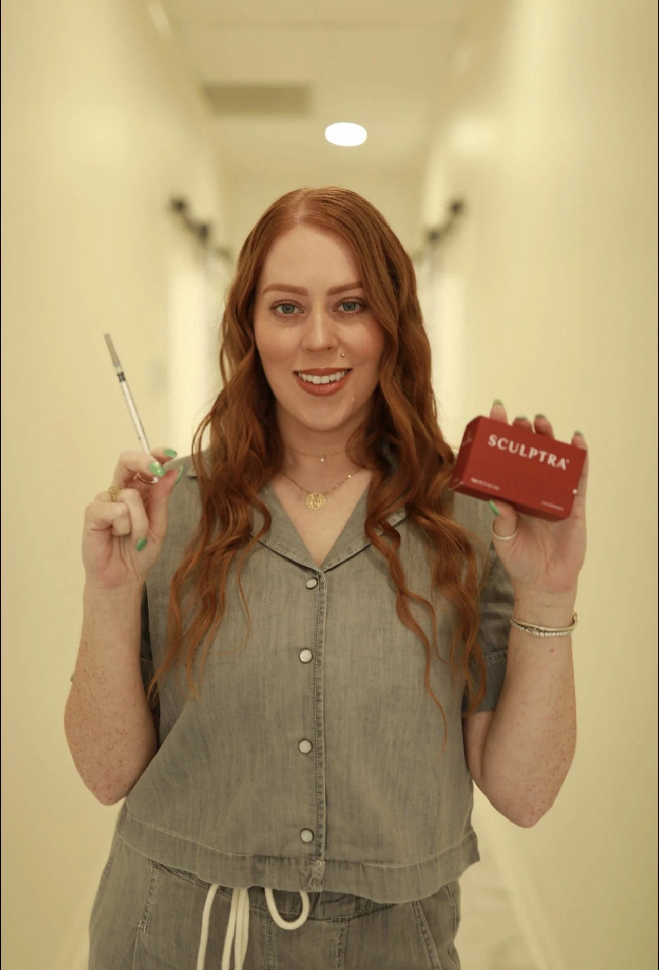 A woman with long red hair holding a syringe in one hand and a red box labeled 'SCULPTRA' in the other, smiling while standing in a corridor.