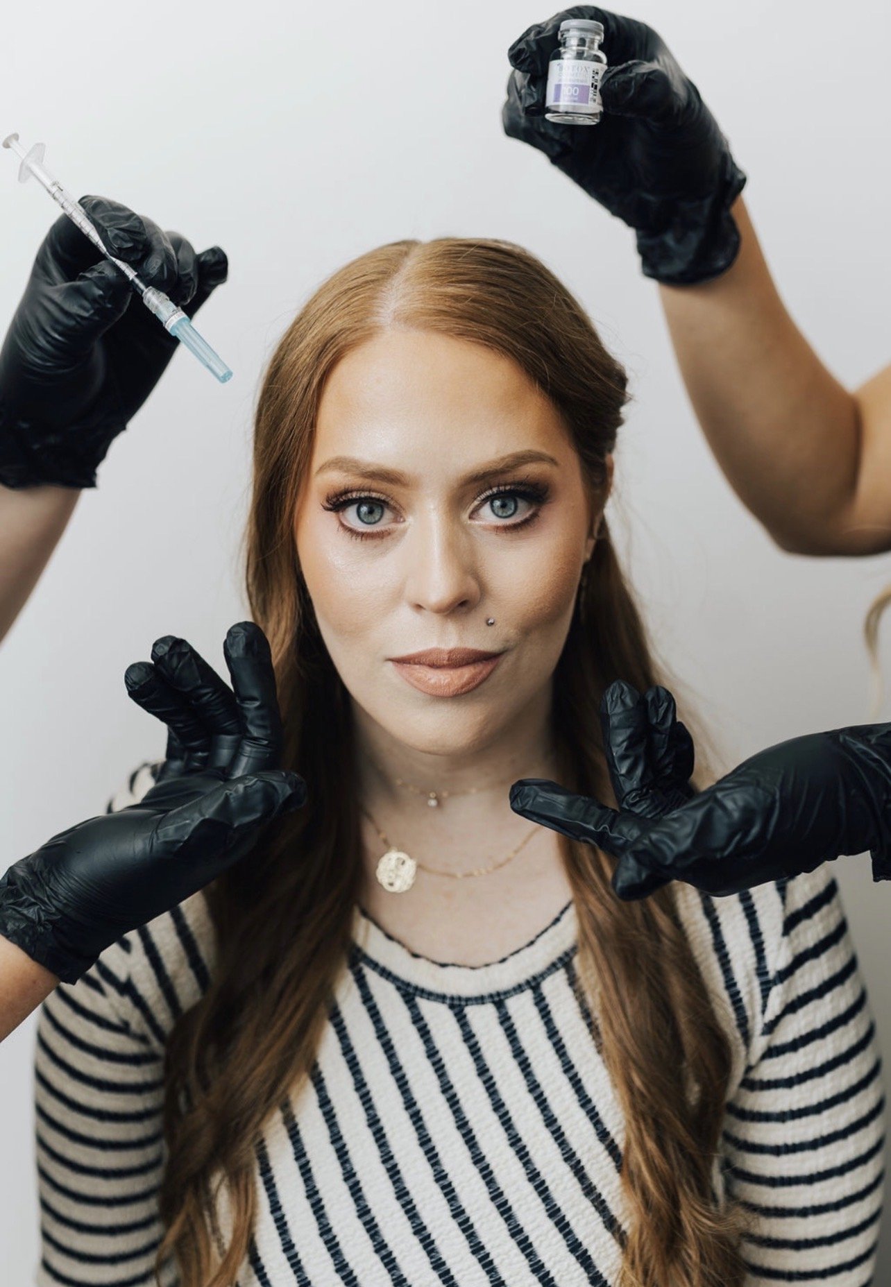 Woman with light skin, blue eyes, and long wavy red hair receiving a cosmetic injection from medical professionals wearing black gloves.