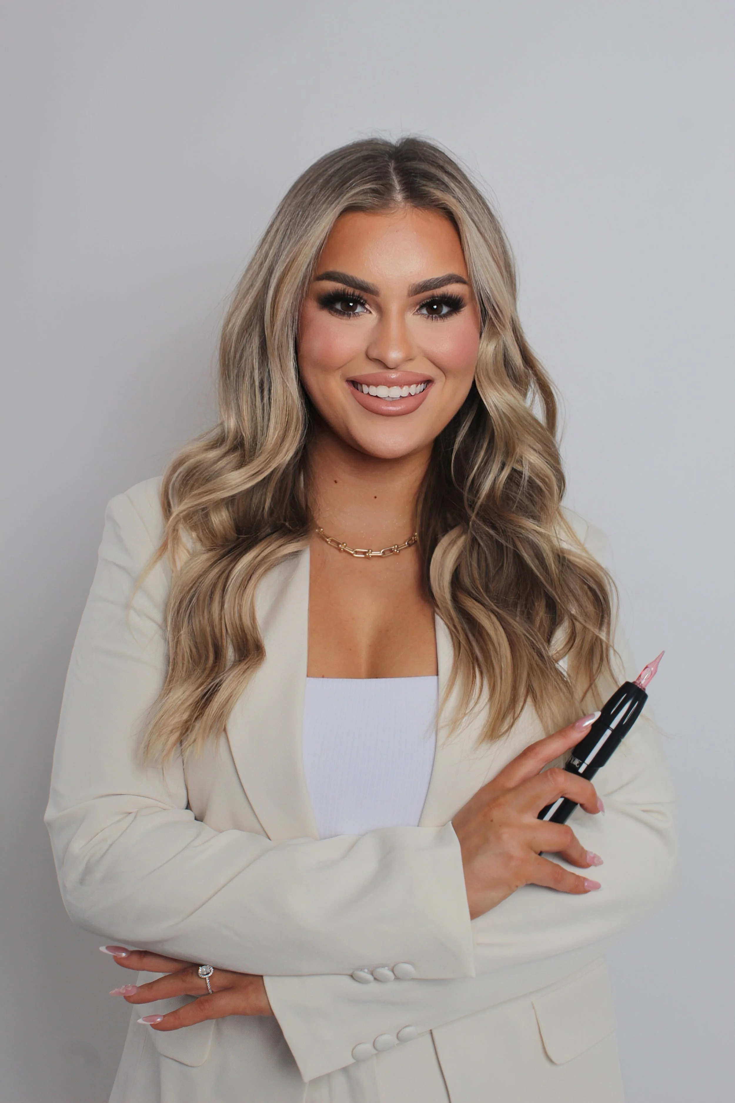 A smiling woman with long blonde wavy hair, wearing a white blazer and top, holding a black marker, against a plain light gray background.