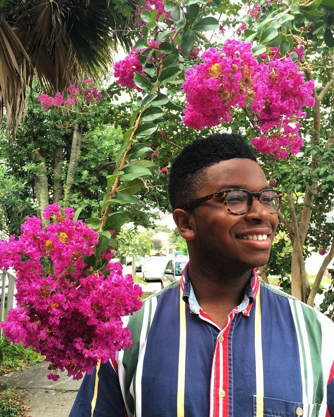 A smiling man with glasses standing outdoors next to a flowering pink crepe myrtle tree.