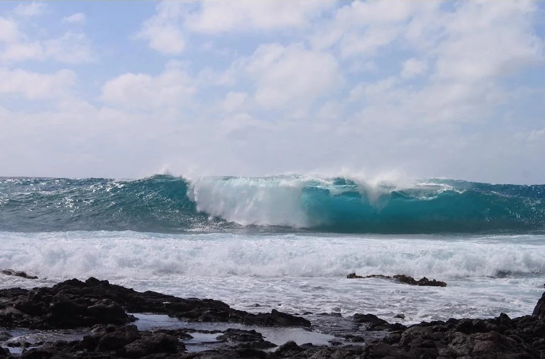 Large ocean wave breaking near a rocky shoreline under a partly cloudy sky.