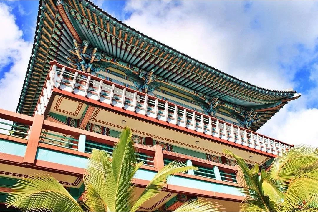 A traditional Asian pagoda-style building with ornate detailing, multiple curved roof tiers, and a balcony with railings, set against a partly cloudy sky with palm trees in the foreground.