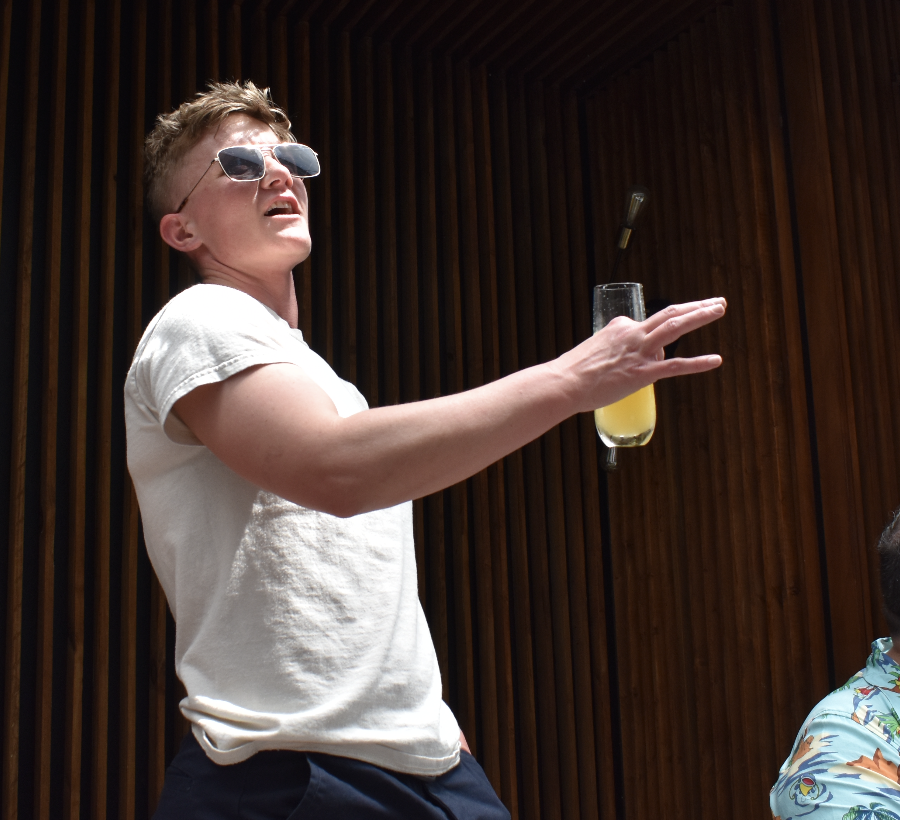 Young man wearing sunglasses and a white t-shirt, gesturing with his hand, with a glass of yellow beverage in mid-air.