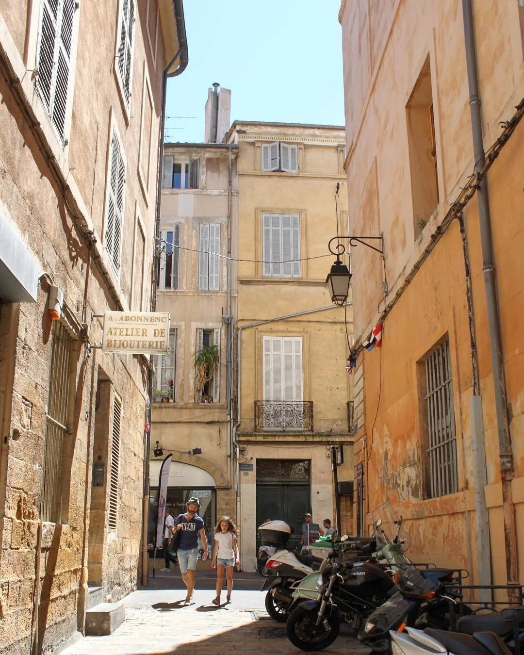 A narrow street in a European city with old yellow and beige buildings. People are walking, and there are parked motorcycles on the sidewalk. A sign reads 'A. Abonnengo Atelier de Bijouterie'.