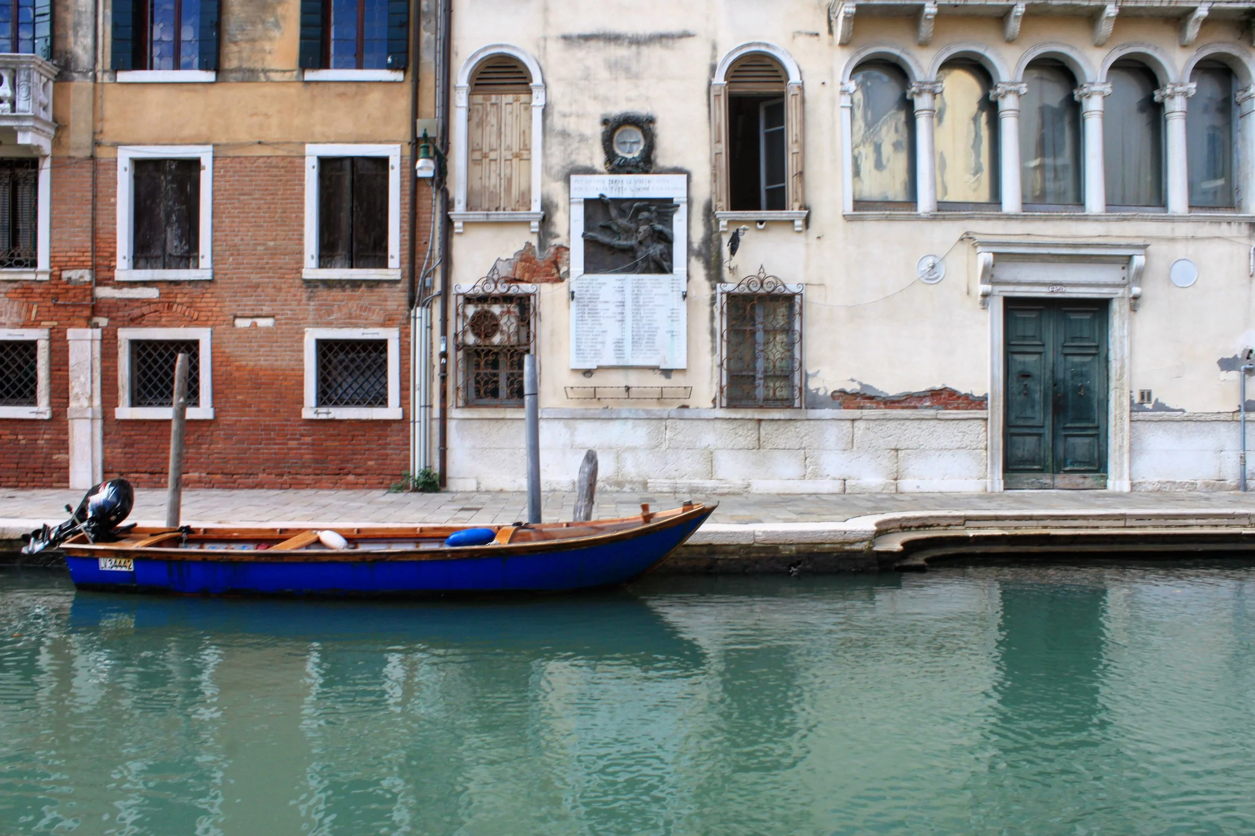 A canal scene with a blue boat floating on green water in front of old, weathered buildings with boarded-up and broken windows in Venice, Italy.