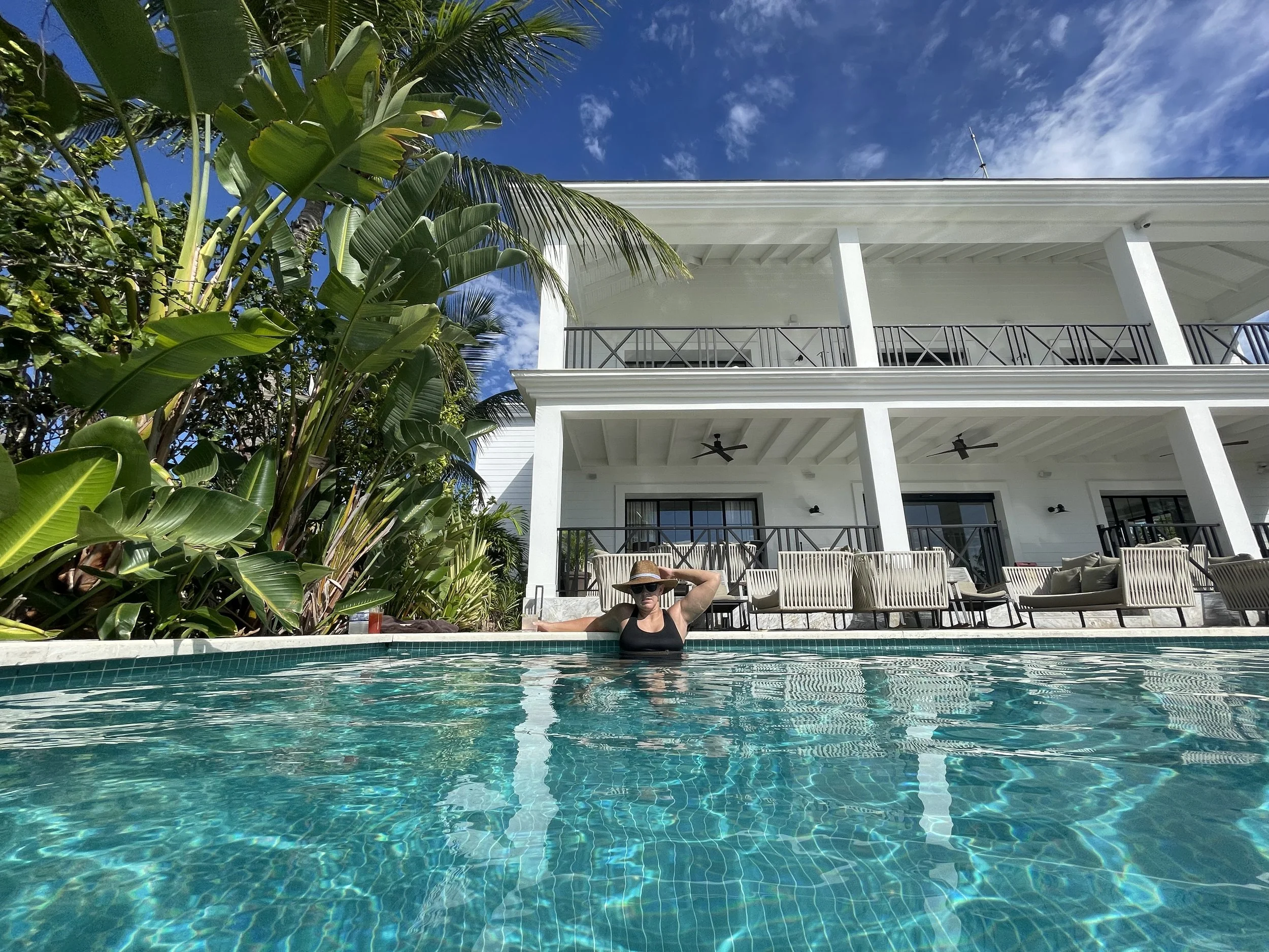 A woman in a black swimsuit and wide-brimmed hat relaxing in a swimming pool, with a white house and tropical plants in the background.