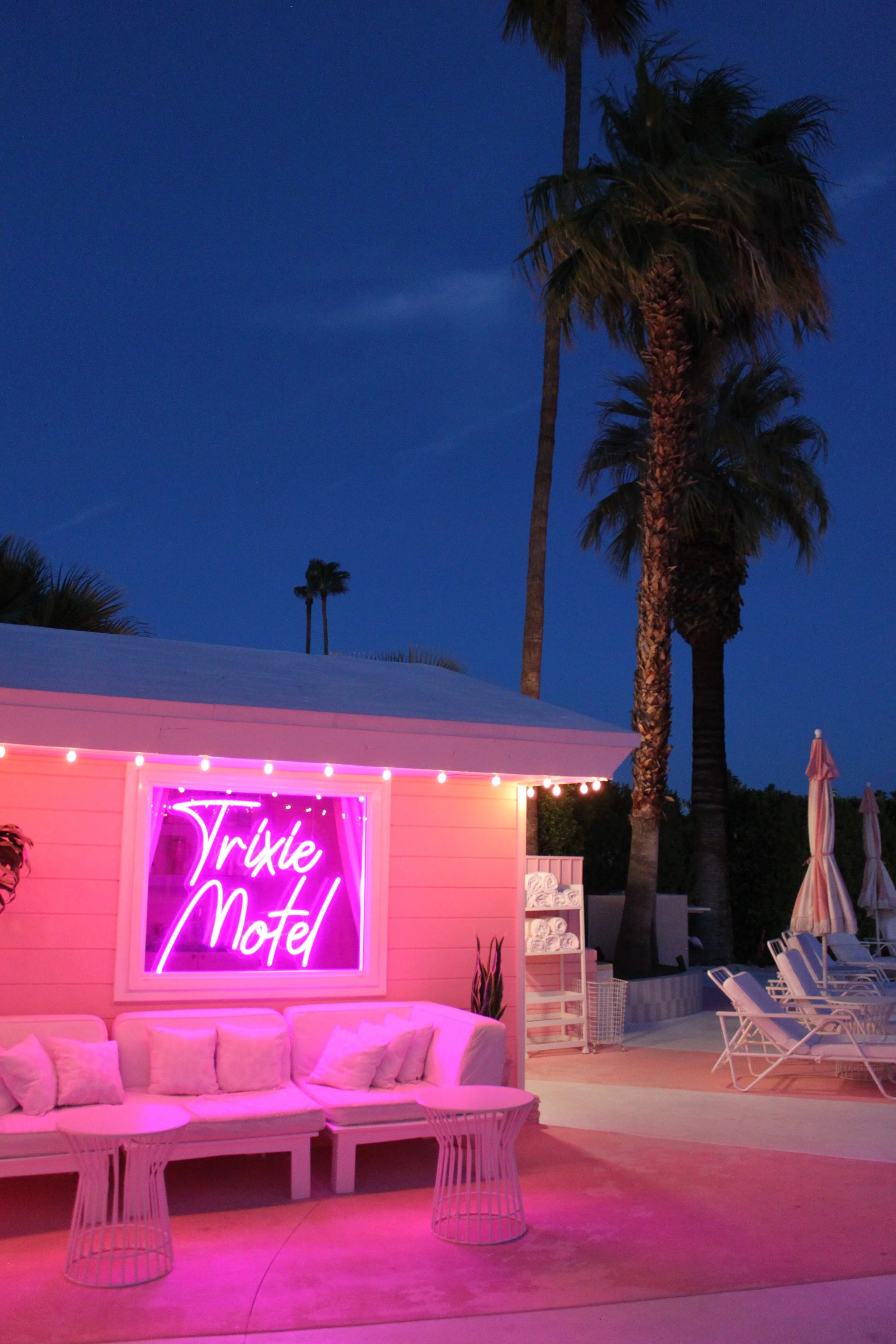 Nighttime view of the Trixie Motel with neon sign, outdoor lounge area with white sofa and pillows, palm trees, and pool chairs with umbrellas.