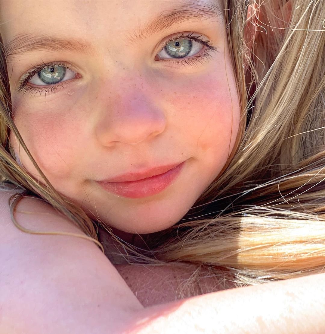 Close-up of a young girl with blue eyes, freckles, and light brown hair, smiling softly.