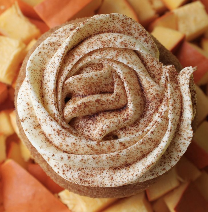 Close-up of a cupcake topped with swirled cream frosting and sprinkled with cinnamon, with chopped fruit in the background.