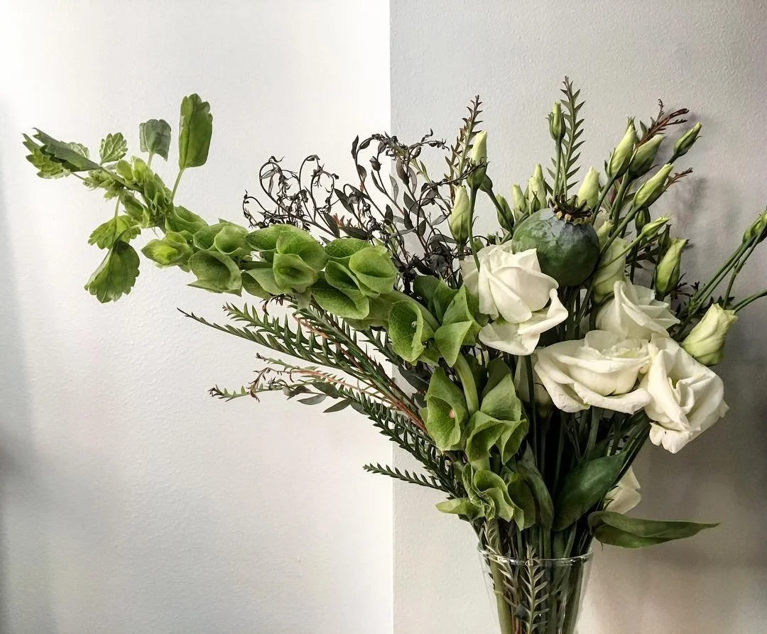 A bouquet of green and white flowers in a clear glass vase against a white and light gray wall.