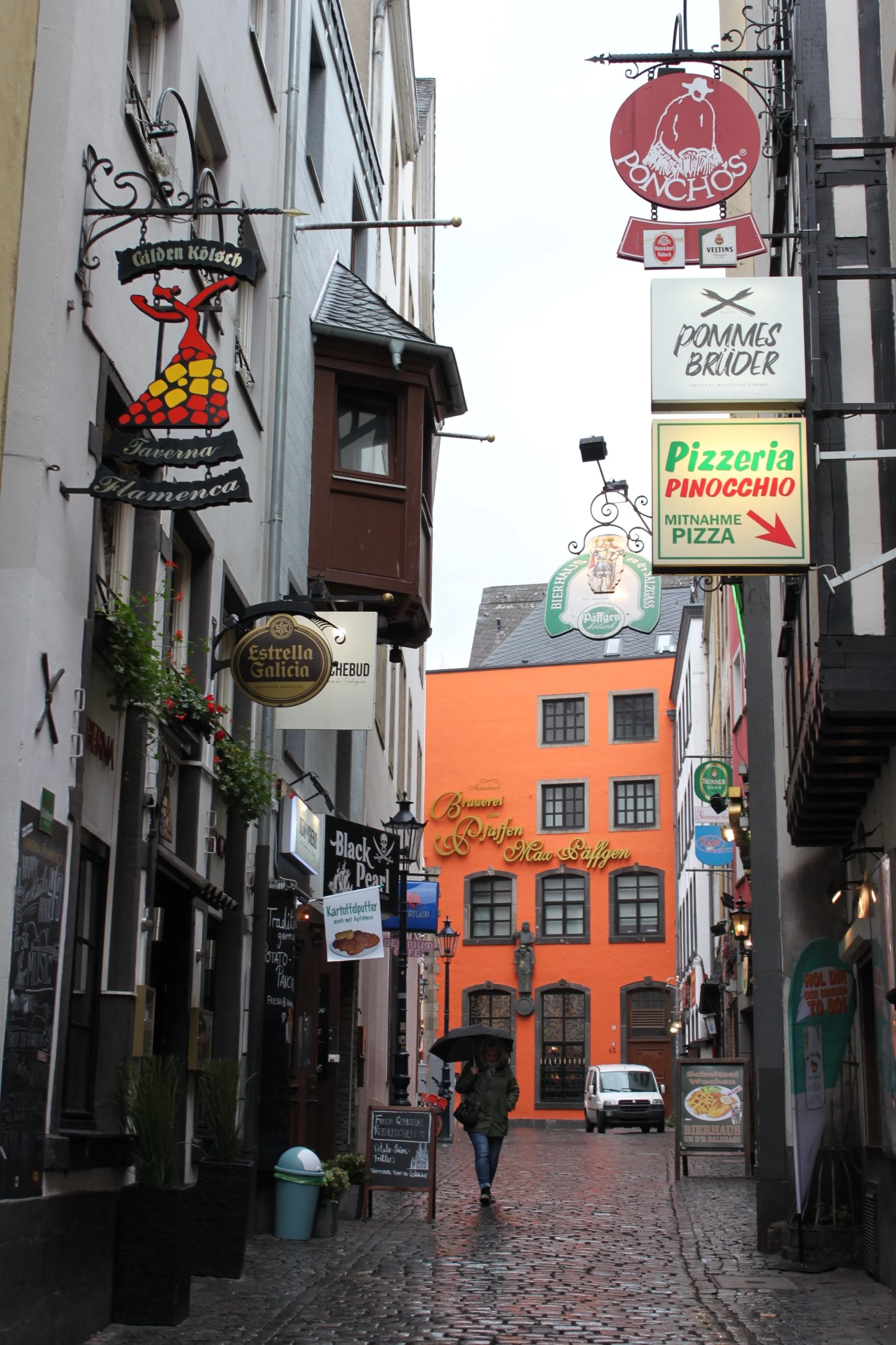 Narrow street with brick pavement, shops and signs on both sides, a person walking with umbrella, closed garage door, orange building in background.