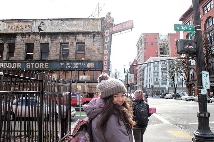A young woman wearing a gray knit hat and purple jacket smiling at the camera on a city street corner with pedestrians and cars, tall buildings, and a large neon sign that says 'PORTLAND OUTDOOR' in the background.