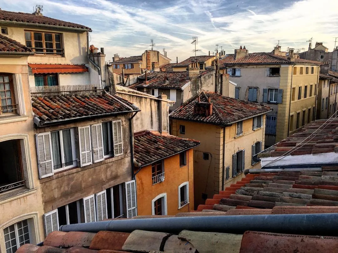 View of a historic European cityscape with closely packed buildings featuring terracotta-tiled roofs, stucco facades, and multiple windows, under a partly cloudy sky.