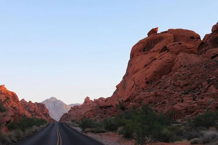 A two-lane desert road running through red rock formations with sparse green bushes, under a clear blue sky.