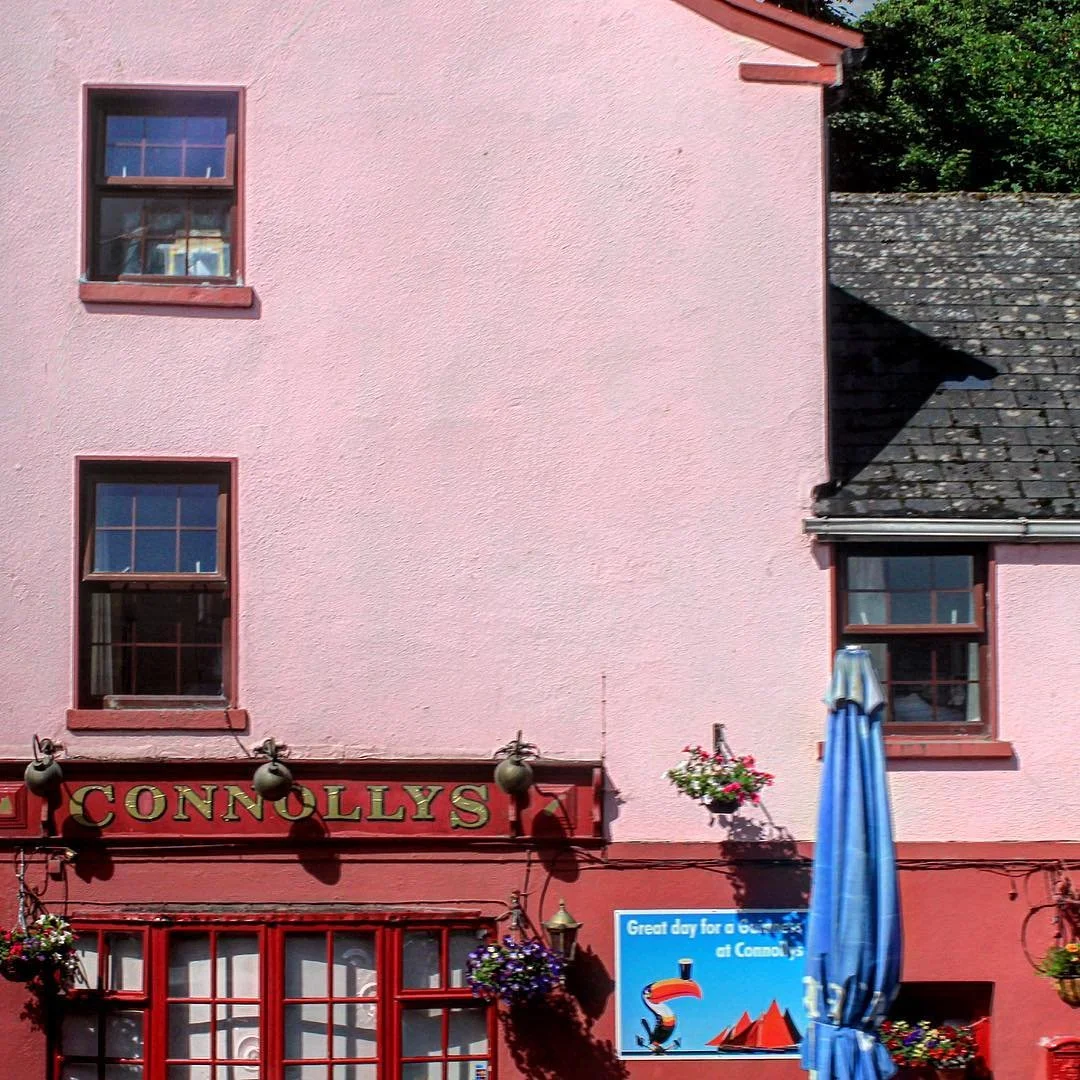 The exterior of a pink building with three windows, flowers hanging outside, and a sign that says 'Connolly's'. There is a large blue umbrella and a decorative toucan poster on the wall.
