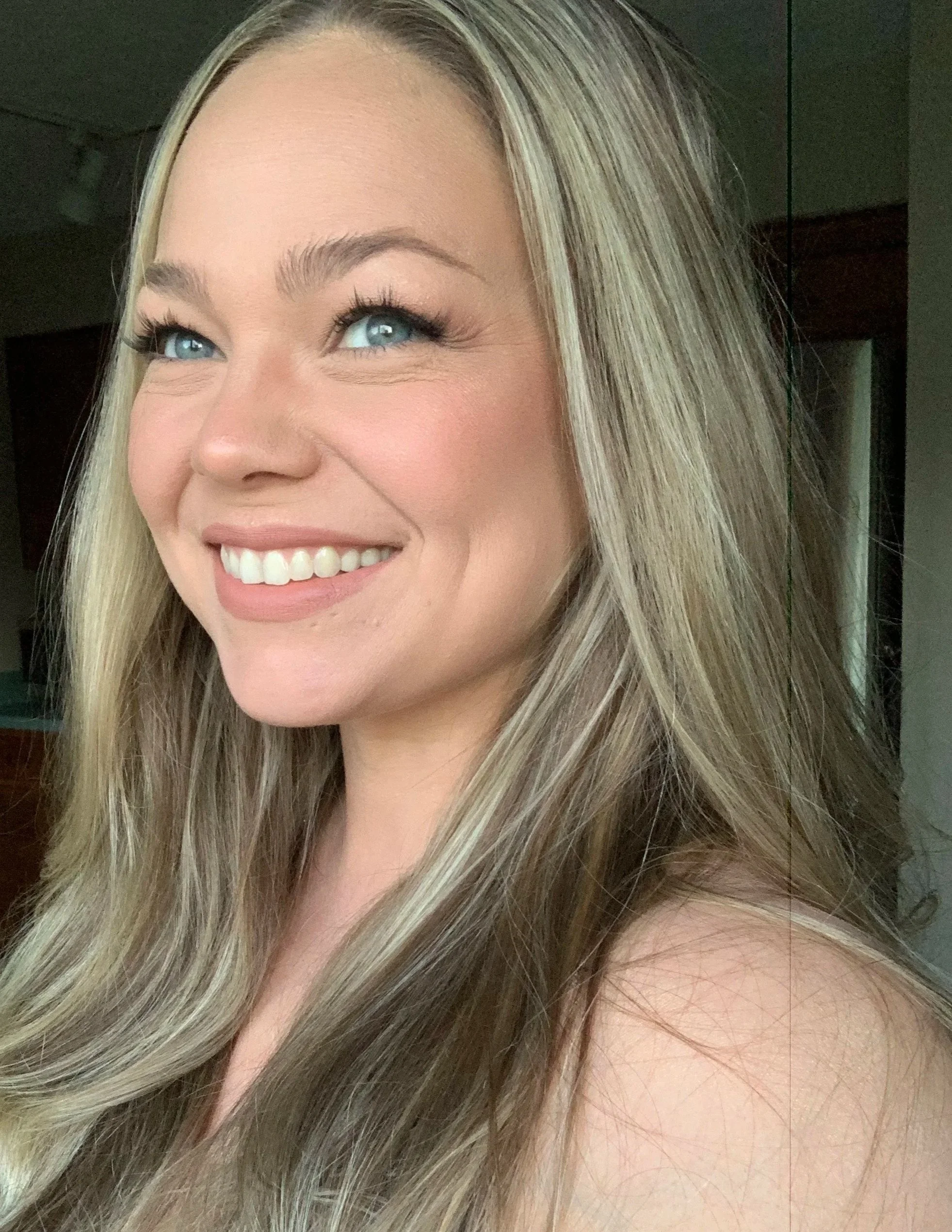 Close-up of a woman with long, blonde hair, blue eyes, and a smile, indoors.