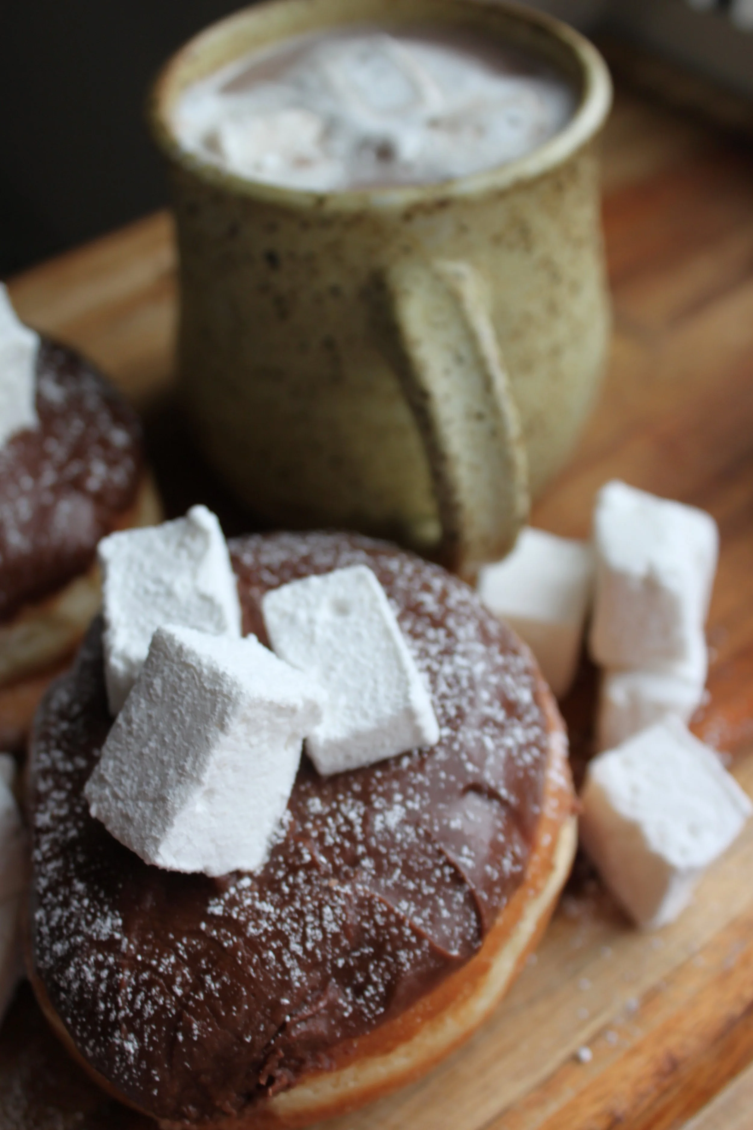 A mug of hot chocolate next to chocolate-glazed donuts topped with powdered sugar and marshmallows on a wooden surface.