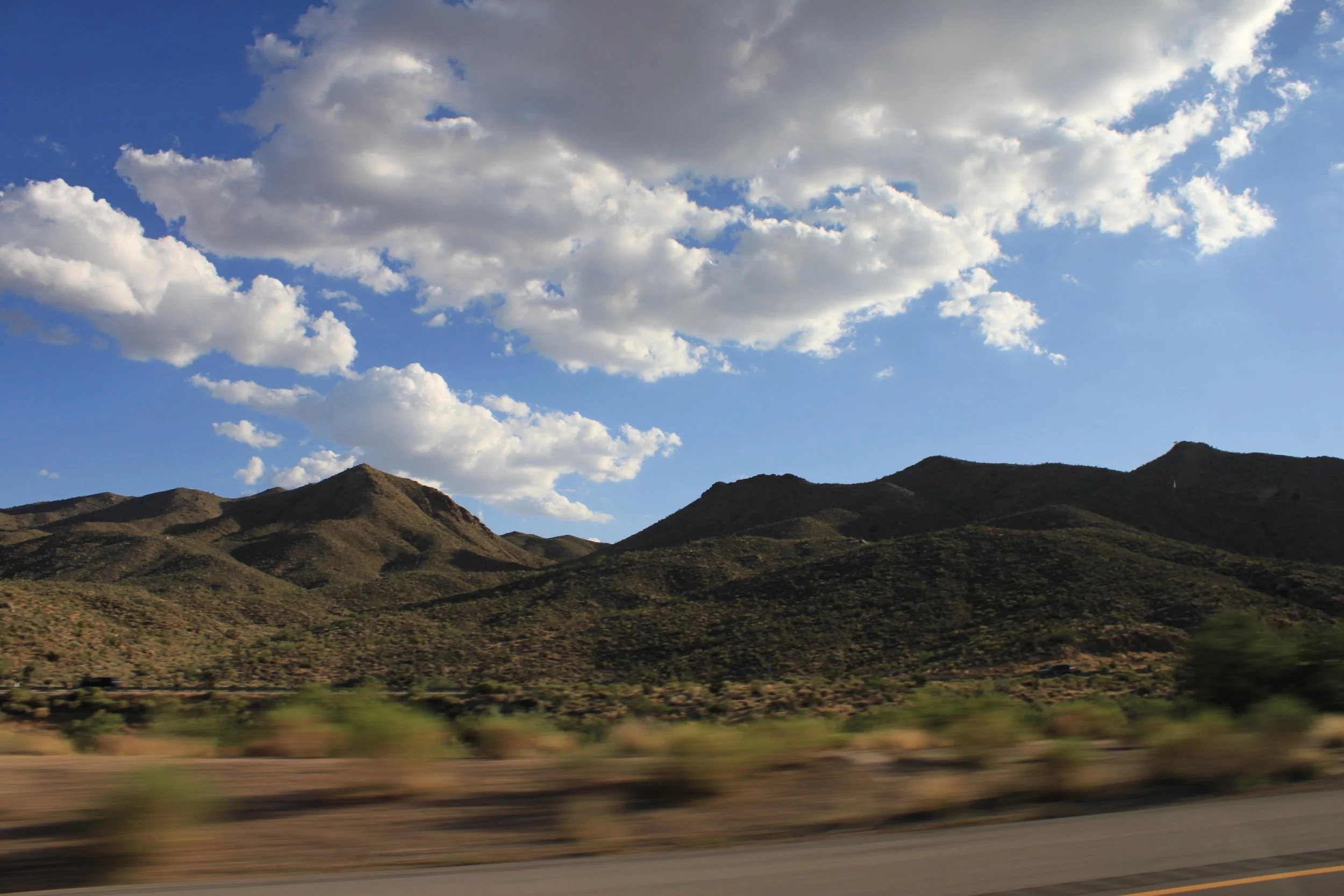 Scenic view of a mountain range under a blue sky with scattered clouds, with a blurred road and desert vegetation in the foreground.
