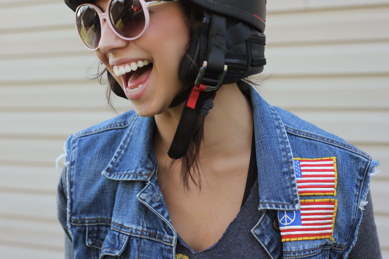 A woman wearing sunglasses, a black helmet, a denim vest with an American flag patch, and a gray shirt, smiling and laughing outdoors.