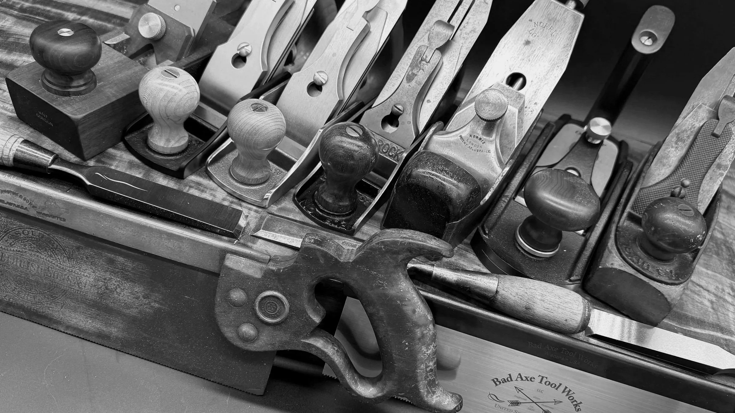 Black and white photo of various hand tools including chisels, a hammer, and a hand plane, arranged on a wooden surface, with a box labeled 'Bad Axe Tool Works' in the background.