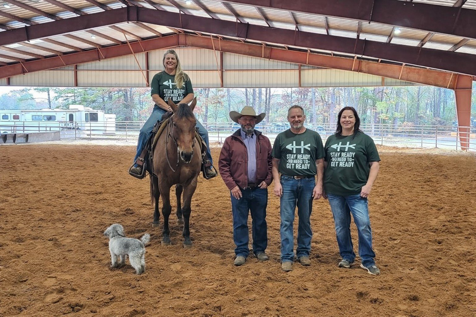 Four people standing inside a horse riding arena, with one person sitting on a brown horse. The rider is a woman with long blonde hair, wearing a green T-shirt. The three individuals on the ground include a man wearing a cowboy hat and maroon jacket, and two others in matching green T-shirts with a white logo and text. A small fluffy dog stands on the ground in front of them. The arena has a dirt floor and a metal roof, with trees visible outside in the background.