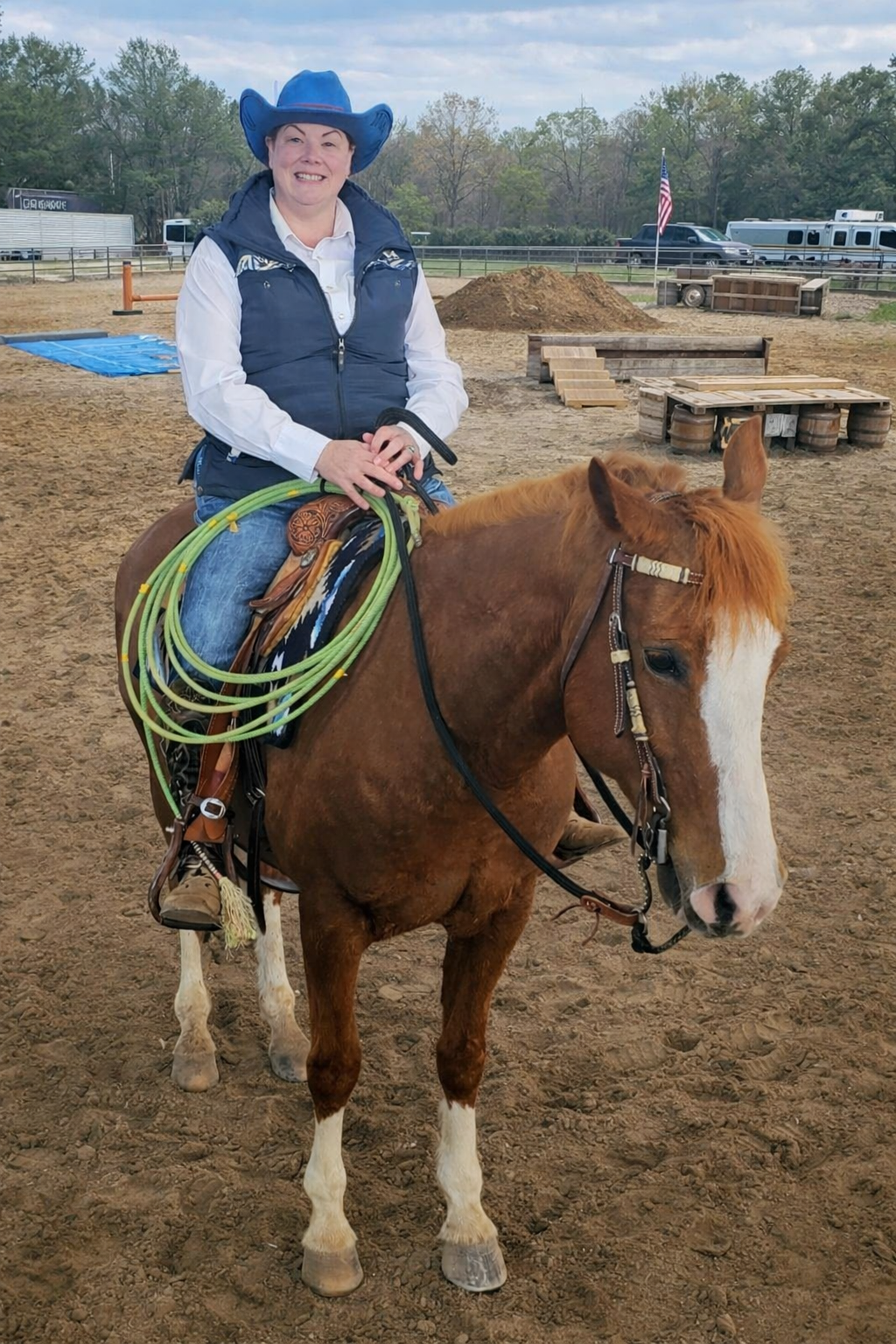 A woman wearing a cowboy hat and vest riding a brown and white horse in an outdoor riding arena, with a trail riding rope and saddle on the horse, and American flags in the background.