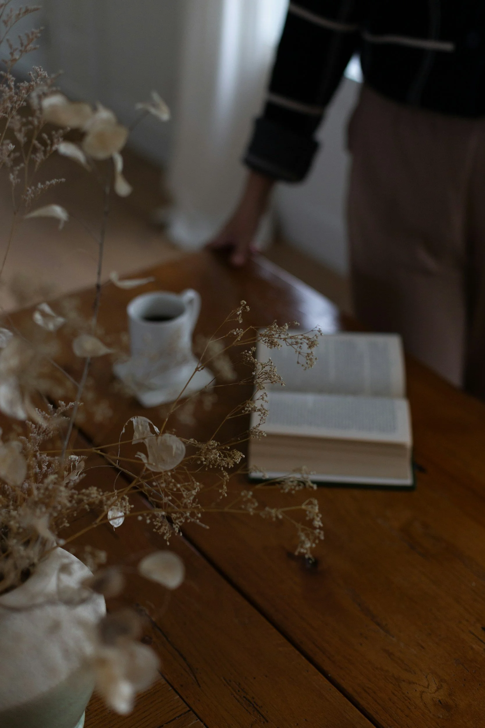 Open book, mug of coffee, and dried plants on a wooden table in a cozy room.