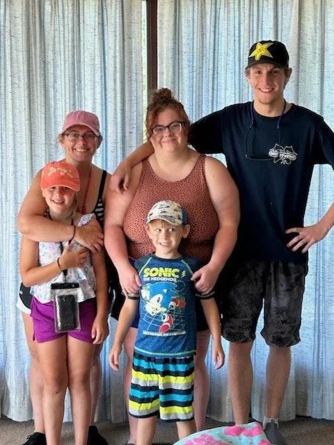 A group of five people, including three children and two adults, standing together indoors in front of a curtain. The children are wearing casual summer clothes and hats. The adults are smiling, with the woman wearing glasses and the man wearing a cap with a star. The group appears happy and close-knit.