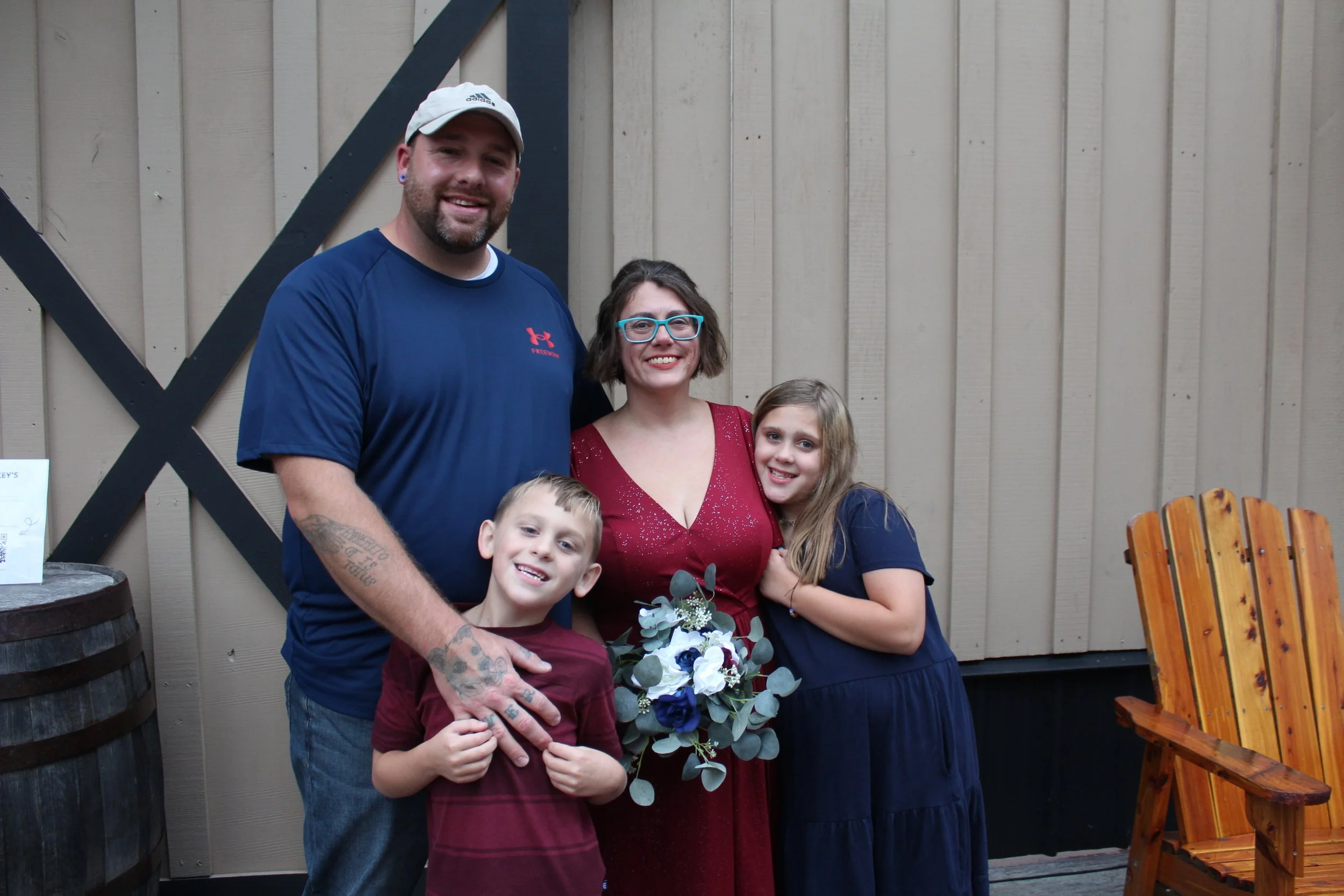 Family of four standing together outdoors, smiling, with a beige painted wooden wall and a wooden chair in the background. The mother holds a bouquet of white and blue flowers with greenery.