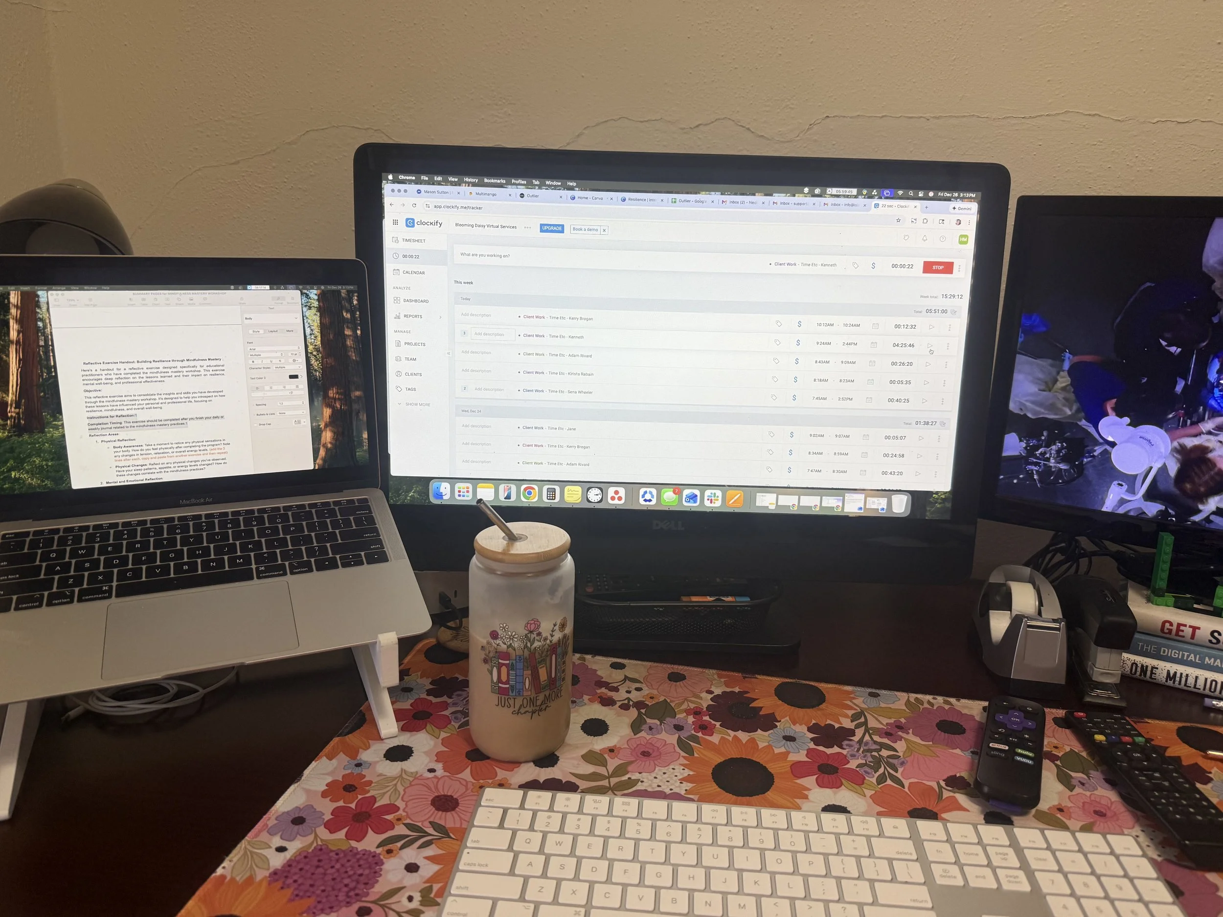 A home office desk with two computer monitors, a MacBook Air, a floral tablecloth, a white keyboard, a remote control, a coffee tumbler with a straw, and various office supplies.