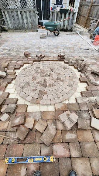 In-progress brick paving with a circular pattern in a backyard, surrounded by loose bricks, wood fencing, and outdoor tools.