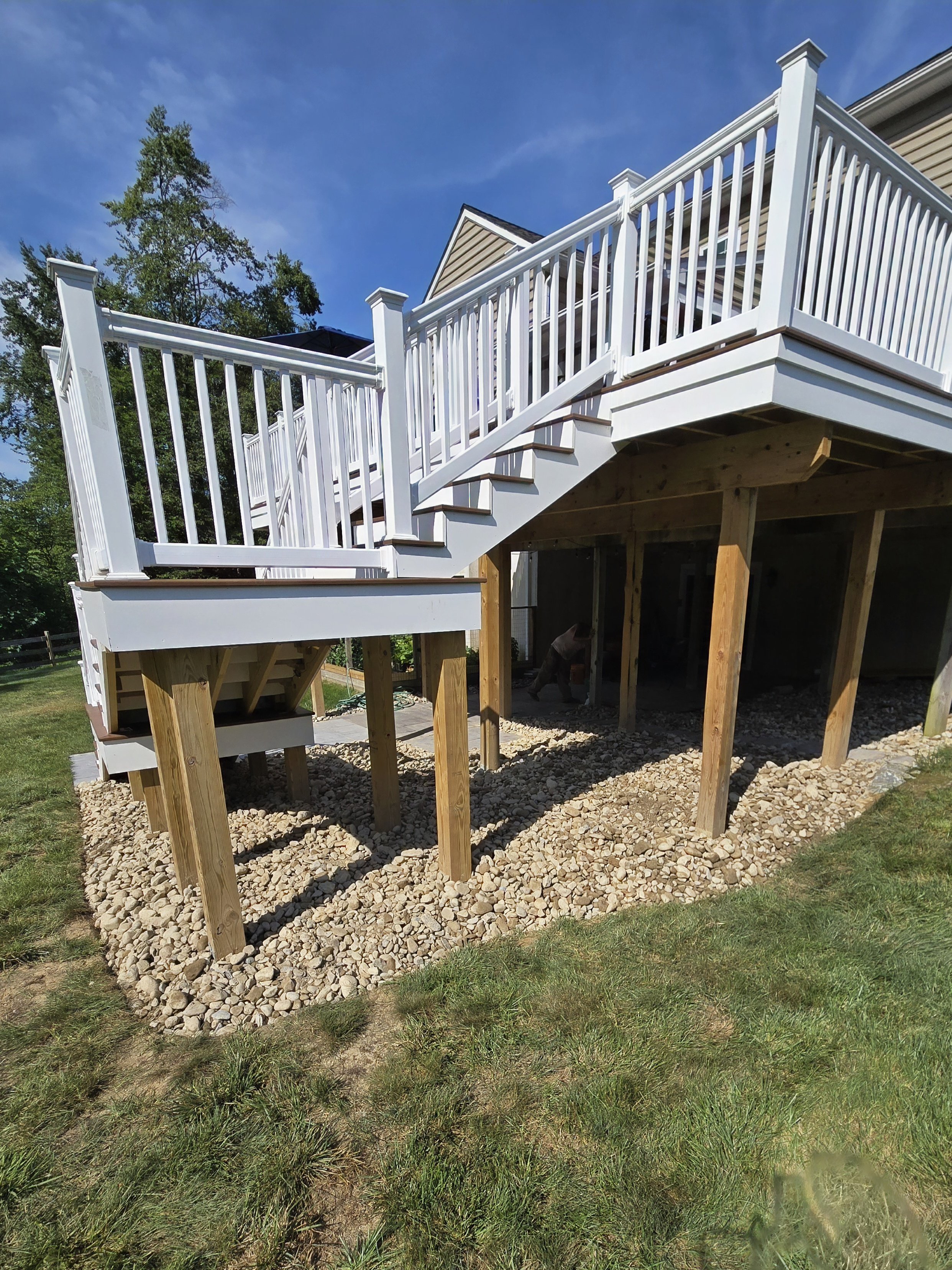 A newly constructed white wooden deck with balcony railing and stairs, supported by untreated wood posts, under a blue sky with trees in the background.