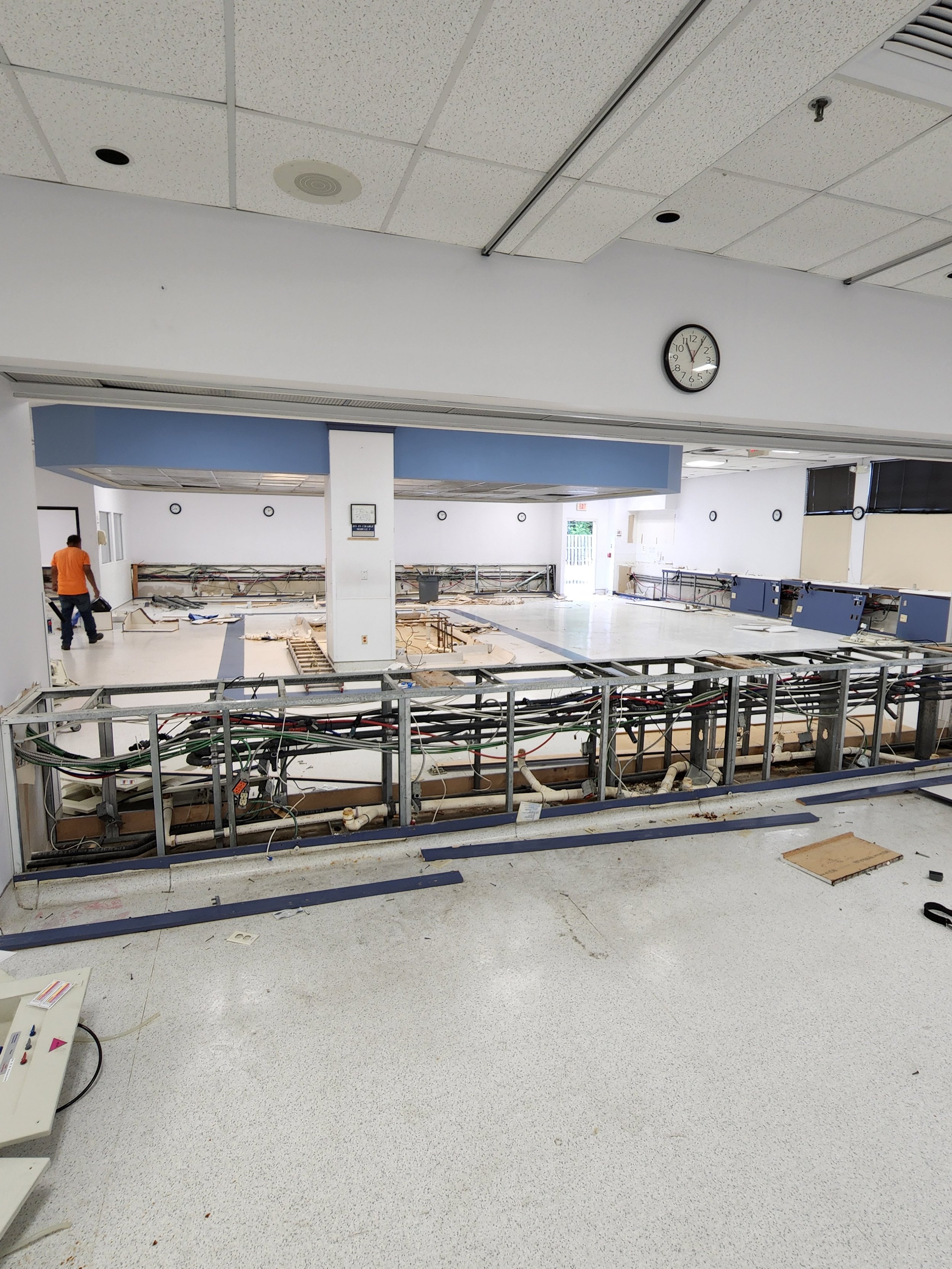 Inside a building under renovation with exposed flooring and wiring, some workers are visible in the background, and the ceiling appears intact with a clock on the wall.