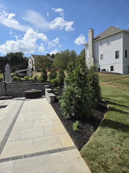 View of a backyard patio with a stone fire pit, surrounded by a landscaped garden with trees, and a white house with multiple windows and a chimney in the background under a partly cloudy sky.