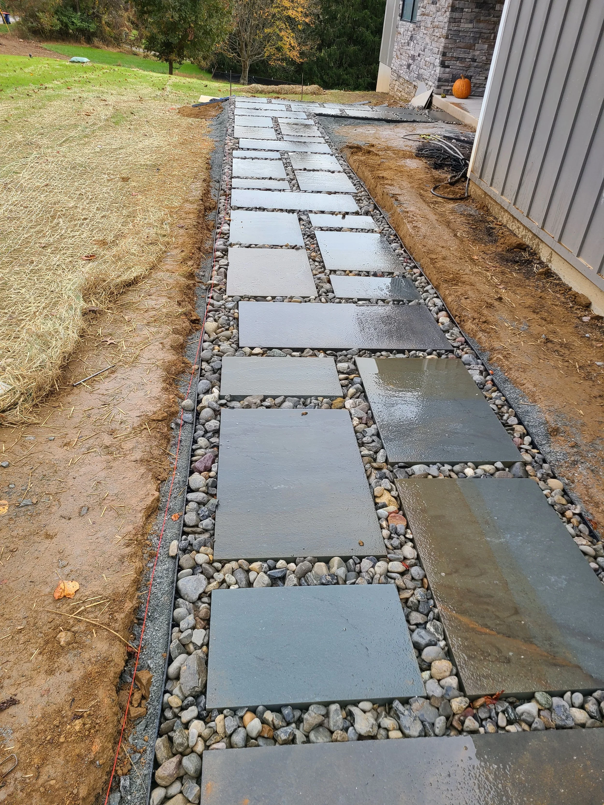 A newly constructed stone pathway with large flat gray stones, surrounded by small rocks, leading from a house to the yard, with a pumpkin on the porch.