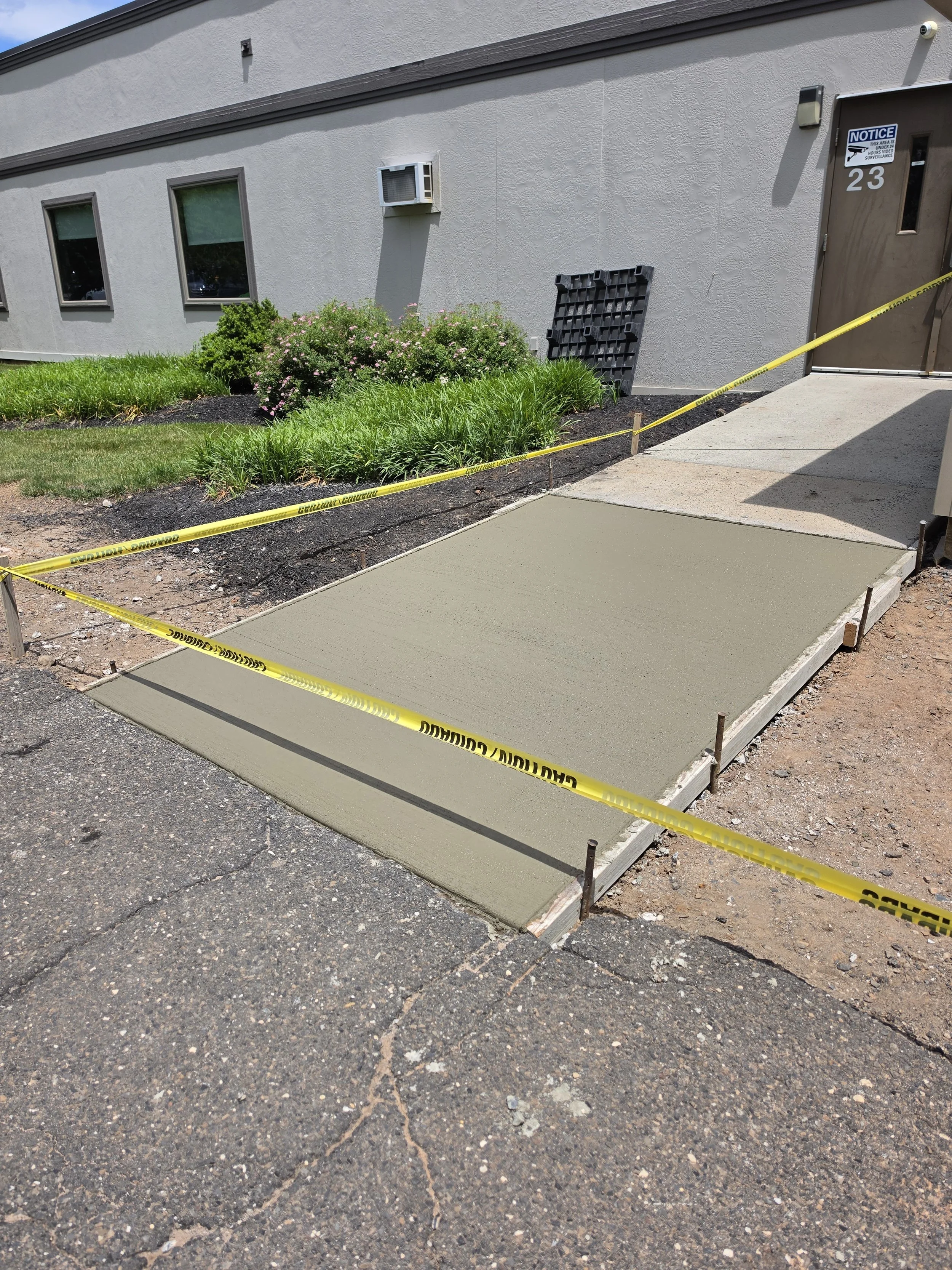 Freshly poured concrete sidewalk in front of a building, blocked off with yellow caution tape, with green plants and a bush nearby.