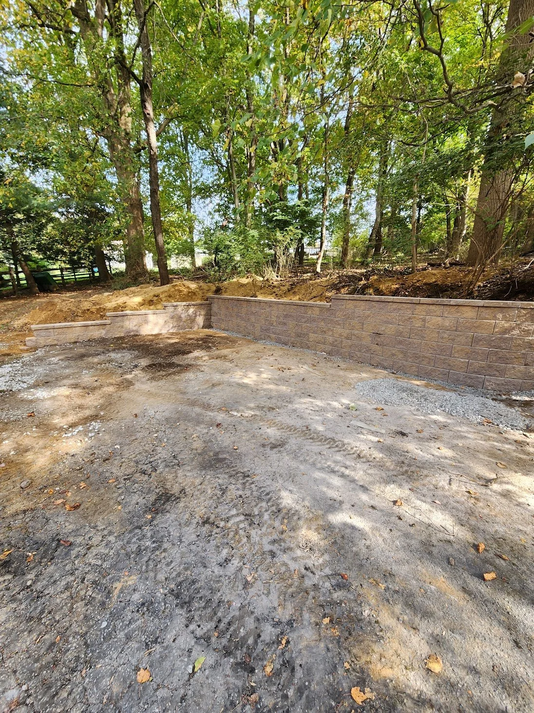 A newly constructed retaining wall made of bricks or blocks, with steps at one end, set against a wooded background, with a cleared dirt surface in the foreground.