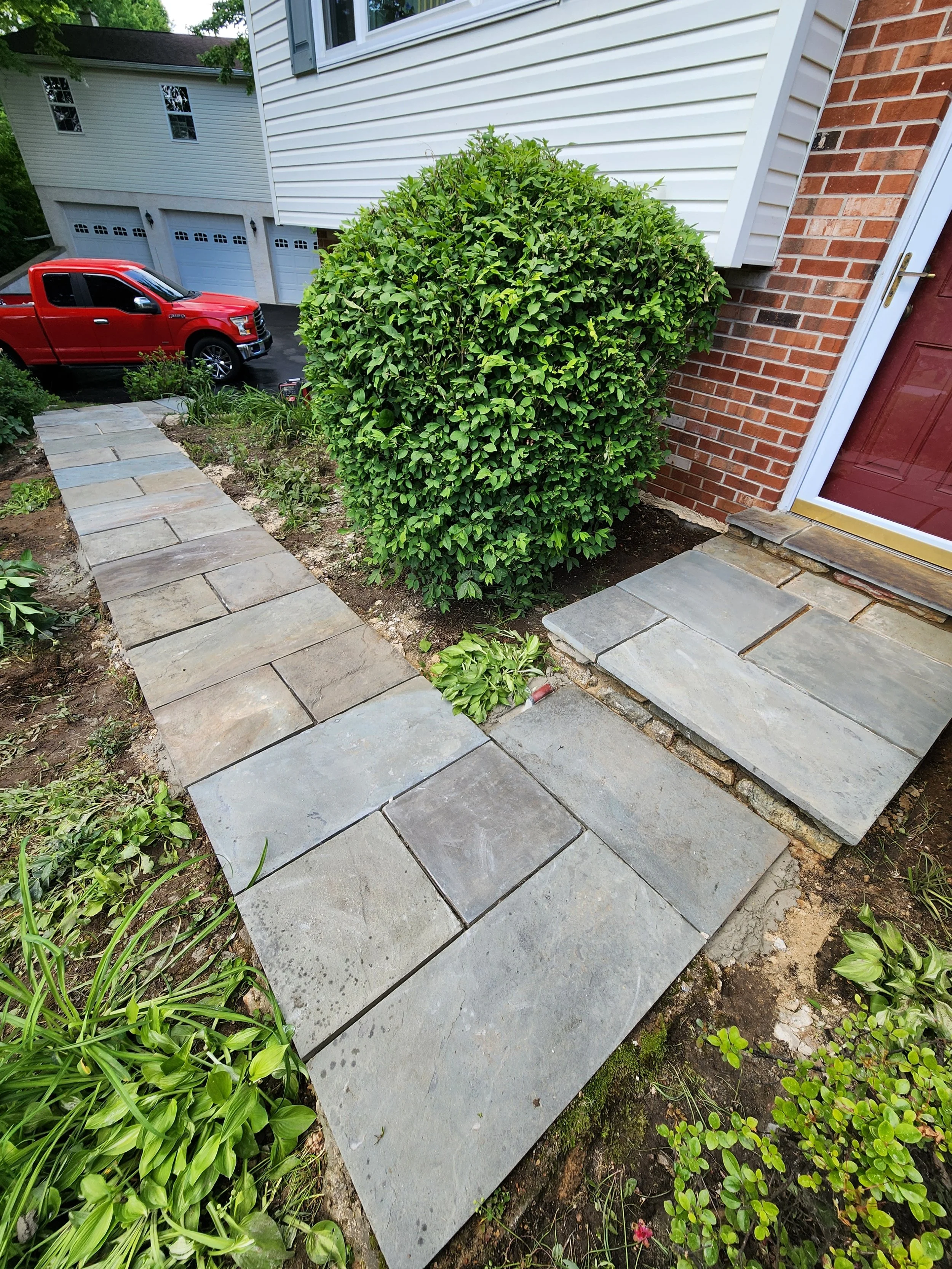 Stone pathway with plants on either side leading to a house with a red door and a large, rounded green shrub in the front yard. A red truck is parked on the street.