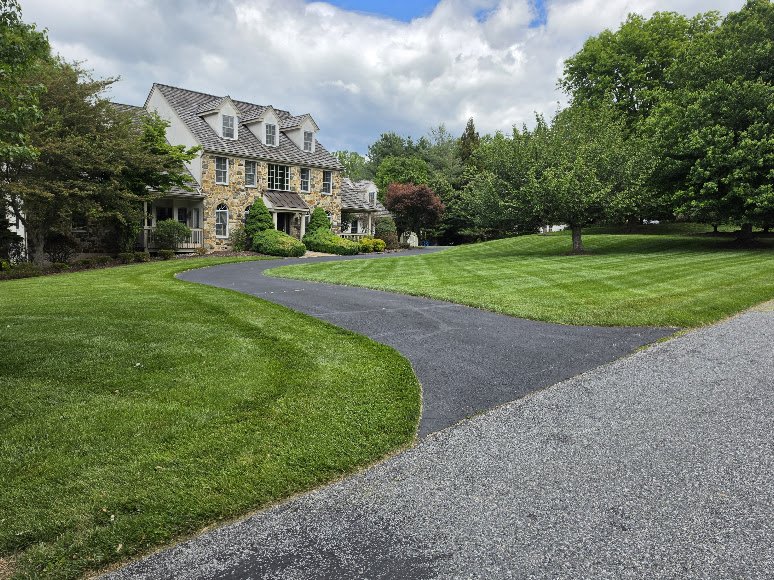 A large house with stone and siding exterior, multiple windows, and a gabled roof, surrounded by well-maintained green lawns and mature trees, with a paved driveway leading to the house, under a partly cloudy sky.