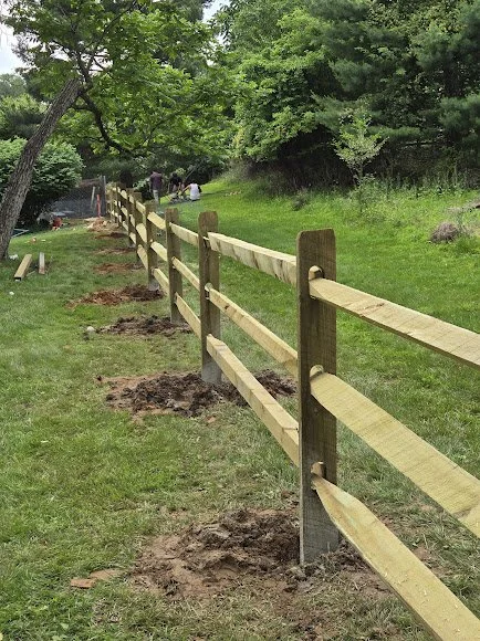 Fence installation with wooden posts and horizontal rails along a grassy area, with people working in the background.