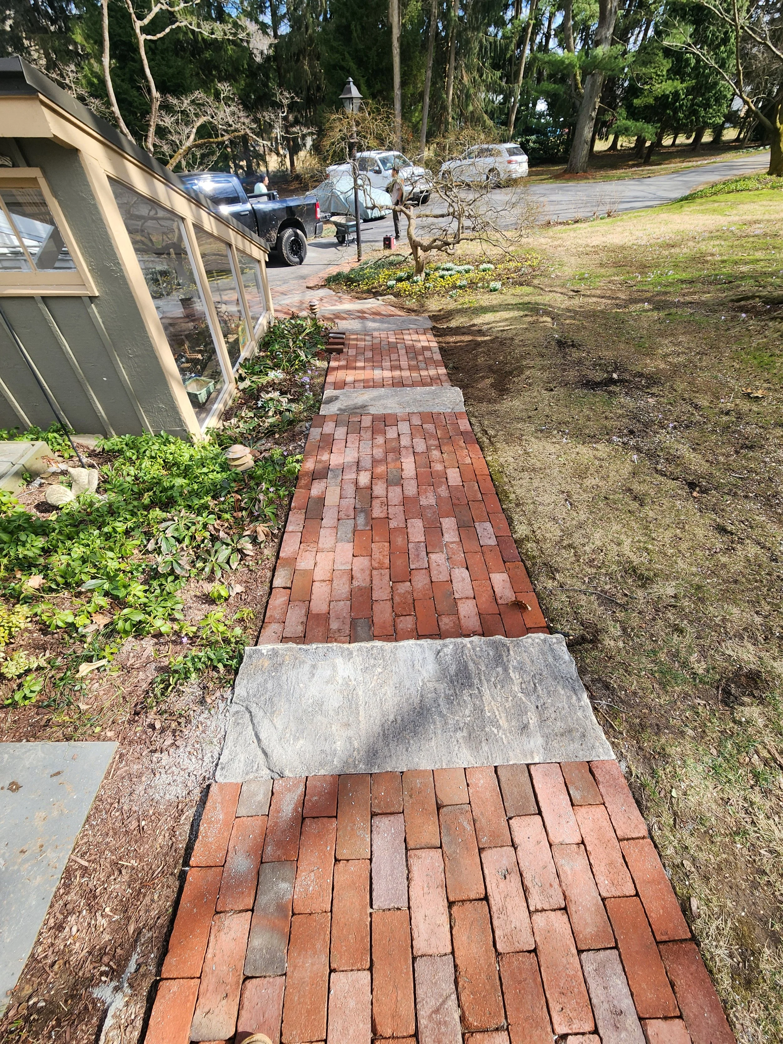 A brick pathway beside a house with a glass windowed sunroom, leading to a flower bed with a small, twisted tree and yellow and white flowers. There are parked cars and trees in the background.