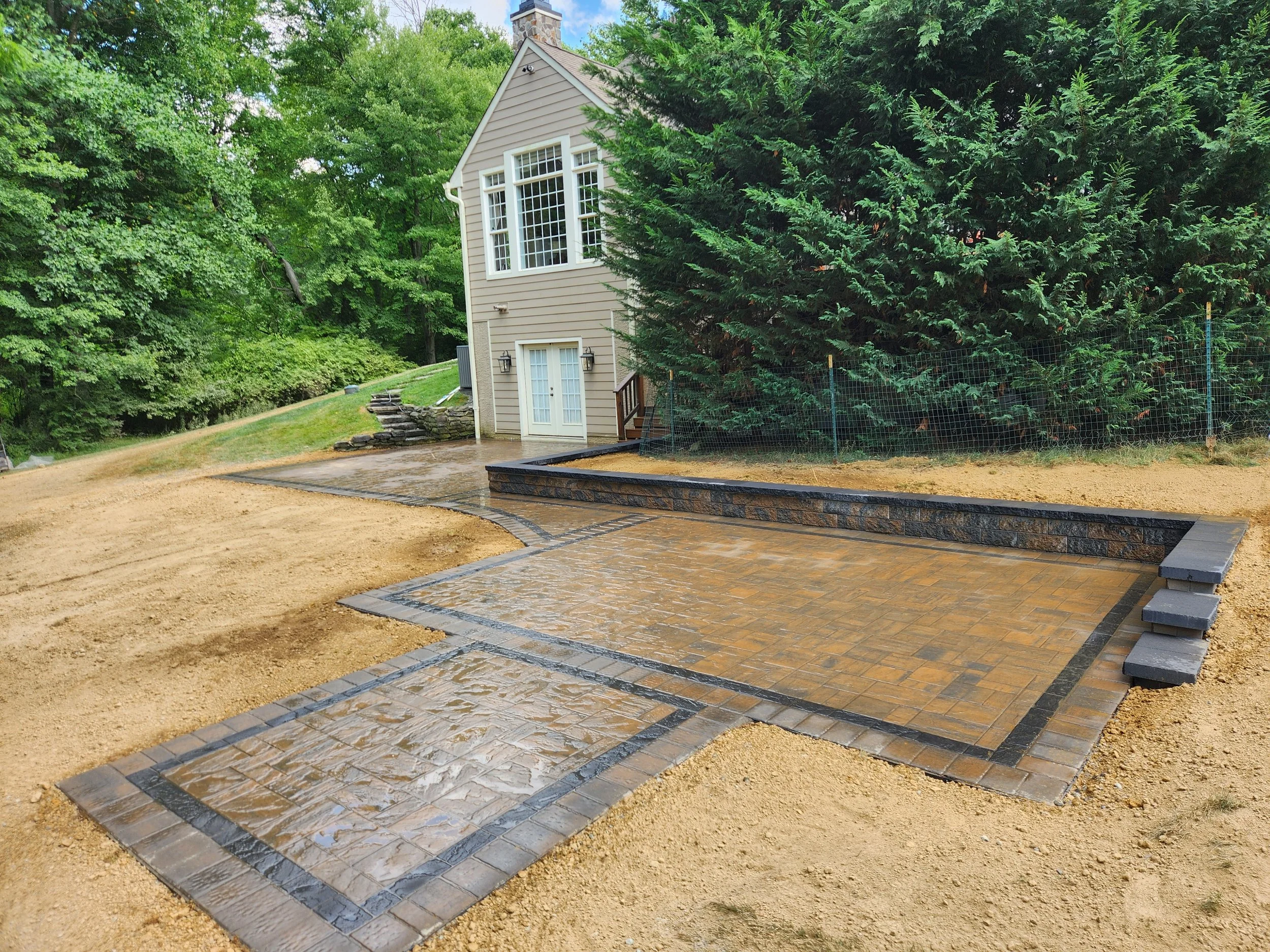 A house with beige siding, large windows, and a small porch surrounded by lush green trees. There is a freshly paved stone and brick pathway leading to the house, with some stone steps nearby. The yard is partly landscaped with a raised stone bed, an