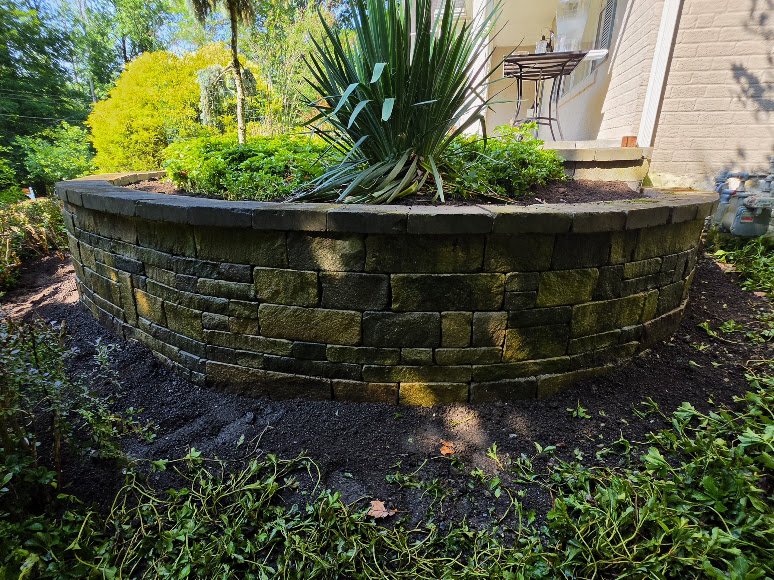 A curved raised garden bed with a stone wall, filled with green plants and a large spiky plant in the center, next to a house with a small outdoor table and chairs.