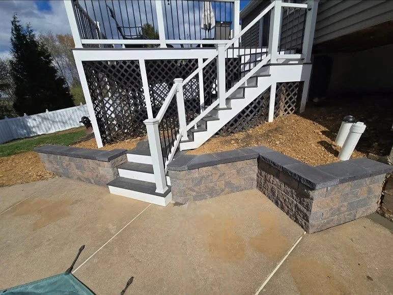 Outdoor stairs with black and white railing leading to a deck, with brick planter walls on either side, and a concrete patio in the foreground.