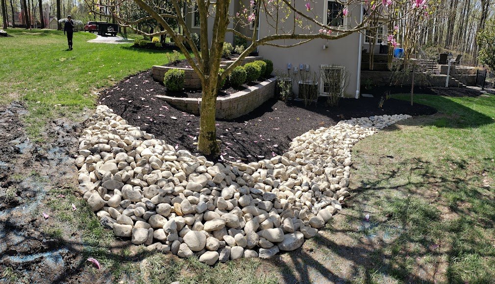 A landscaped backyard with a blooming tree, a gravel pathway with white stones, a small garden bed with dark mulch, and a house with a deck in the background.