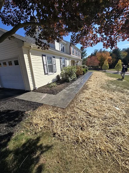 Side view of a light yellow house with a driveway, surrounded by trees with fall foliage, and a person walking on the sidewalk.